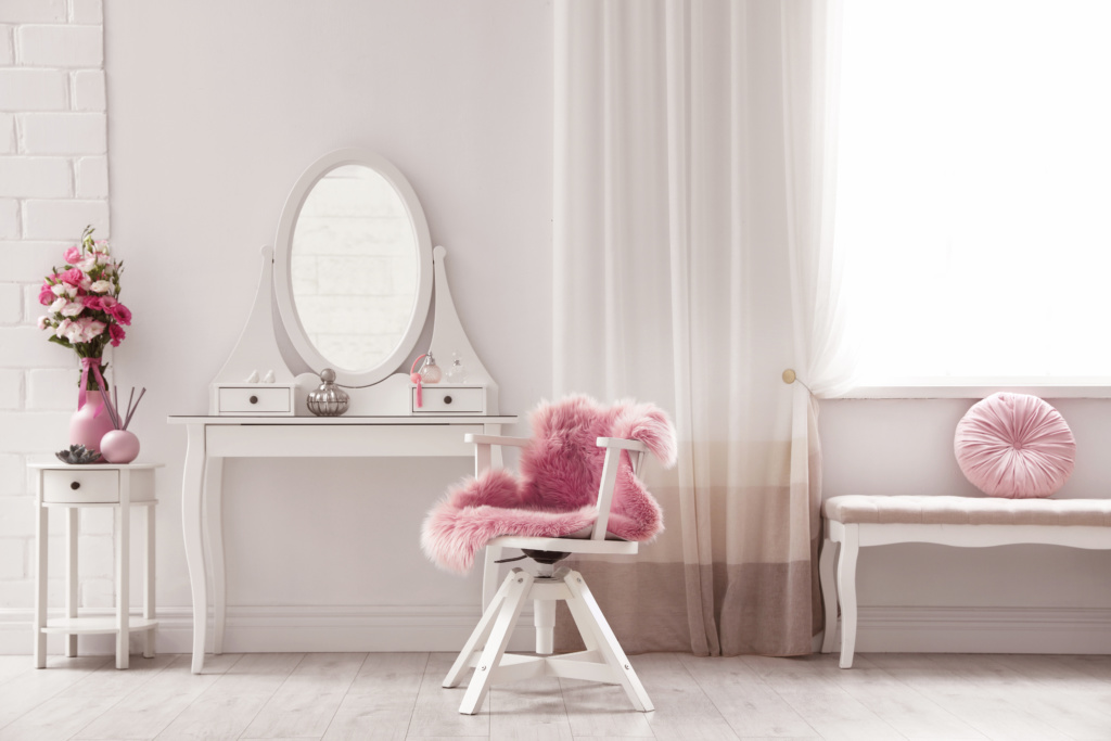 Decoração de quarto feminino com uma penteadeira retrô com espelho, cadeira e um tecido de pele na cor rosa, além de ter um ambiente bem iluminado.
