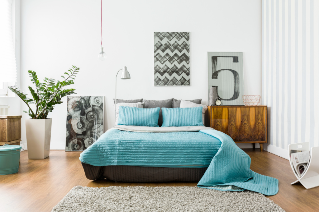 Decoração de com uma cama de casal com enxoval azul, móveis no tom de madeira, vaso com planta e um tapete na cor cinza.